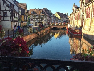 Ein malerischer Kanal in Colmar, Frankreich, mit bunten Fachwerkhäusern, einer Brücke und Blumen unter blauem Himmel.