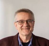 Porträt eines lächelnden Mannes mit kurzen grauen Haaren und Brille, der ein kariertes Hemd und eine dunkelrote Jacke trägt.