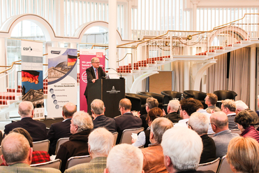 Mann spricht auf einem Podium vor Publikum bei einer Konferenz über Sicherheitspolitik, mit deutscher Flagge und Treppe im Hintergrund.