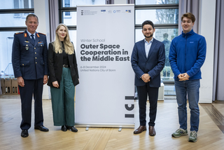 Vier Personen, darunter ein Mann in Uniform, posieren vor einem Banner der 'Winter School Outer Space Cooperation'.