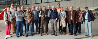 Eine Gruppe von etwa 20 lächelnden, leger gekleideten Erwachsenen, meist Senioren, die in einem Flugzeughangar posieren.