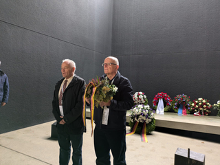 Zwei Männer bei einer Gedenkfeier mit Blumenkränzen und Deutschlandbändern vor einer dunklen Wand.