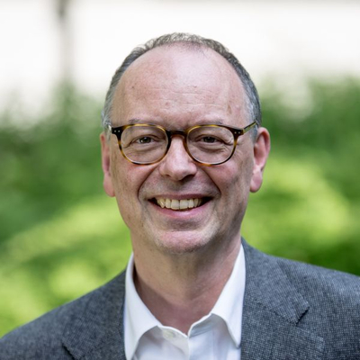 Porträt eines lächelnden Mannes mit Brille, kurzen grauen Haaren und grauer Jacke vor unscharfem grünem Hintergrund.