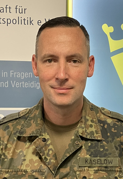 Ein Mann in deutscher Militäruniform mit KASELOW-Namensschild und Deutschlandflagge steht vor einer Wand mit Organisationstext.