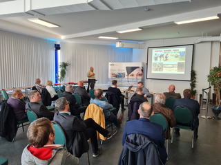 Gruppe von Männern bei einer Präsentation mit einem Redner am Pult und projizierten Folien auf einer Leinwand.