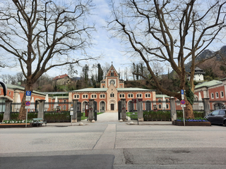 Ein prächtiges Backsteingebäude mit zentralem Uhrturm und Bogenfenstern, gerahmt von zwei großen kahlen Bäumen an einem bewölkten Tag.