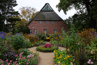 Ein traditionelles rotes Backsteinhaus mit steilem Reetdach und weißen Fenstern, umgeben von einem bunten Garten unter bedecktem Himmel.