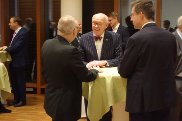 Männer in Anzügen unterhalten sich an Stehtischen bei einer Veranstaltung. Ein Mann mit Fliege lächelt in der Mitte.