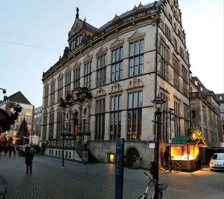 Historisches Bremer Rathaus bei Dämmerung, mit beleuchtetem Weihnachtsmarktstand auf dem Kopfsteinpflasterplatz.
