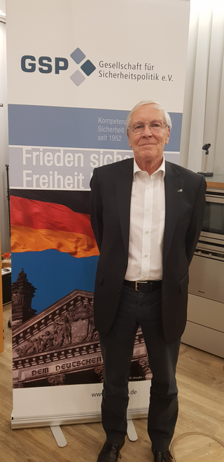 Älterer Mann im Anzug vor einem GSP-Banner mit 'Frieden Sicherheit Freiheit' und deutscher Flagge/Reichstag.