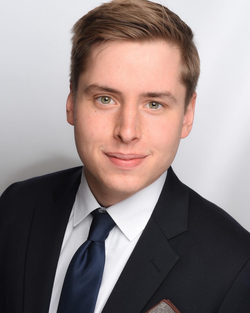 Porträt eines jungen Mannes mit hellbraunen Haaren, grünen Augen, dunklem Anzug, weißem Hemd und blauer Krawatte, lächelt leicht.