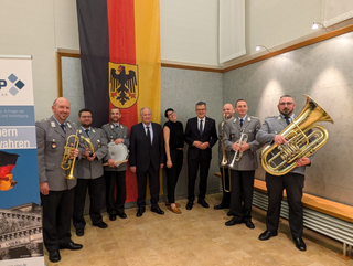 Militärmusiker mit Blasinstrumenten und Trommel, sowie Zivilisten, posieren lächelnd vor der Bundesflagge.