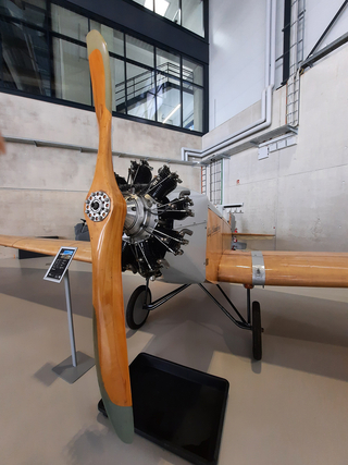 Historischer Motorsegler mit großem Holzpropeller und freiliegendem Sternmotor in einer modernen Ausstellungshalle.