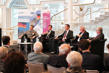 Podiumsdiskussion mit zivilen und militärischen Rednern, von Publikum beobachtet, in hellem Konferenzraum mit Bannern.