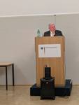 Ein älterer Mann in Anzug spricht an einem Rednerpult mit dem Logo des Bundessprachenamtes. Wasserflasche und Mikrofon sind zu sehen.