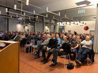 Ein großes Publikum sitzt in einem hellen Raum, lauscht einer Präsentation, ein Rednerpult und Projektor sind sichtbar.