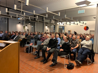 Ein großes Publikum sitzt in einem hellen Raum, lauscht einer Präsentation, ein Rednerpult und Projektor sind sichtbar.