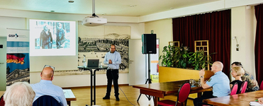 Ein Referent präsentiert vor Zuhörern, mit einer historischen Projektion und einem GSP-Banner 'Frieden sichern Freiheit bewahren'.