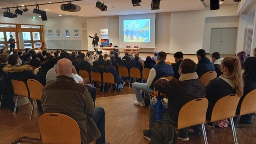 Ein Redner spricht vor einem großen Publikum in einem Saal, dahinter ein bunter Cartoon auf einer Leinwand projiziert.