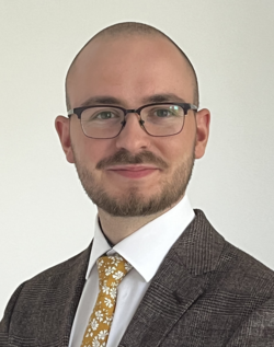 Porträt eines lächelnden Mannes mit Bart, Brille, braunem karierten Sakko und gelber geblümter Krawatte.