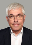 Porträt eines lächelnden Mannes mit kurzen grauen Haaren und Brille, der einen dunklen Anzug und ein weißes Hemd vor grauem Hintergrund trägt.