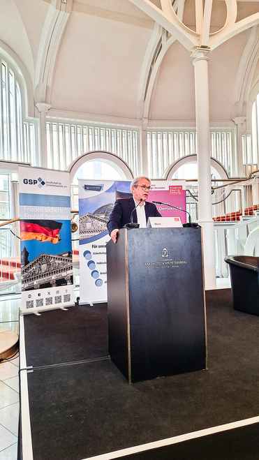 Redner Maier spricht in einem prächtigen Saal am Pult. Im Hintergrund Banner der GSP und Friedrich-Ebert-Stiftung.