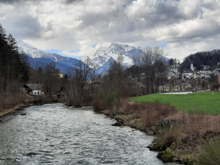 Ein breiter Fluss fließt durch ein Tal mit einer Stadt am Berghang und schneebedeckten Bergen unter bewölktem Himmel.