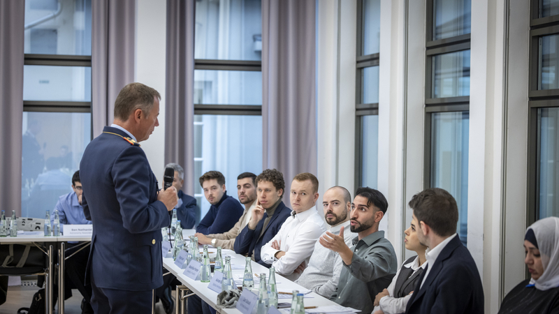 Mann in Uniform spricht vor einer Gruppe junger Erwachsener an einem Konferenztisch mit Namensschildern und Wasserflaschen.