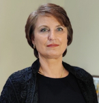 Porträt einer lächelnden Frau mit kurzen braunen Haaren, blauen Augen, rotem Lippenstift und schwarzer bestickter Jacke.