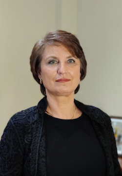 Porträt einer lächelnden Frau mit kurzen braunen Haaren, blauen Augen, rotem Lippenstift und schwarzer bestickter Jacke.