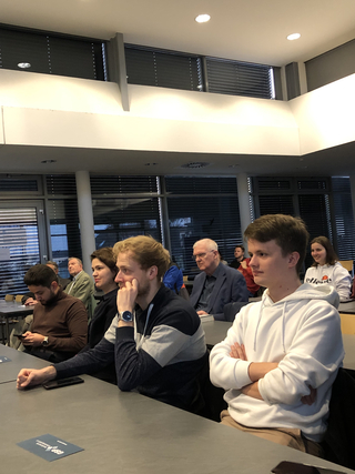 Verschiedene Personen, jung und alt, aufmerksam an Tischen sitzend bei einer Indoor-Veranstaltung oder einem Vortrag.
