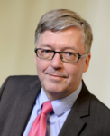 Porträt eines mittelalten Mannes mit grauem Haar, Brille, dunklem Anzug, hellblauem Hemd und pinker Krawatte, lächelnd.