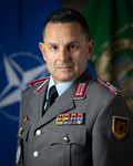 Porträt eines deutschen Bundeswehr-Offiziers in Uniform mit Medaillen vor unscharfem NATO-Flaggenhintergrund.