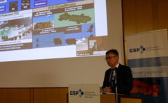 Oberst Asmus spricht bei einer GSP-Veranstaltung, dahinter eine Präsentation zu militärischen Expeditionssystemen.