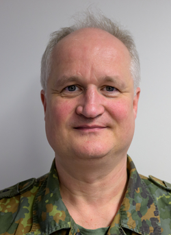 Ein Mann mittleren Alters mit grauem Haar in Tarnuniform lächelt sanft in die Kamera vor einem schlichten Hintergrund.