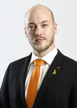 Kahlköpfiger Mann mit Bart im schwarzen Anzug, orangefarbener Krawatte, Deutschlandflagge und gelber Schleife am Revers.