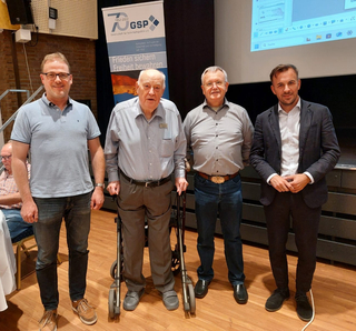 Vier Männer, darunter ein älterer Herr mit Rollator, posieren vor einem Banner und einer Leinwand.