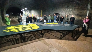 Führung in einem dunklen Tunnel: Gruppe betrachtet Modell von Bahngleisen und -weichen auf einem Tisch, eine Frau erklärt.