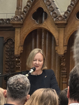Eine blonde Frau mit dunklem Oberteil spricht mit geschlossenen Augen in ein Mikrofon von einer verzierten Holzkanzel zu Publikum.