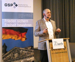 Ein Mann spricht auf GSP-Veranstaltung. Banner: dt. Flagge, Reichstag, Slogan "Frieden sichern Freiheit bewahren".