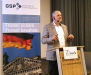 Ein Mann spricht auf GSP-Veranstaltung. Banner: dt. Flagge, Reichstag, Slogan "Frieden sichern Freiheit bewahren".