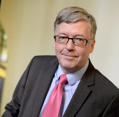 Lächelnder Mann mit grauem Haar, Brille, dunklem Anzug und pinker Krawatte.