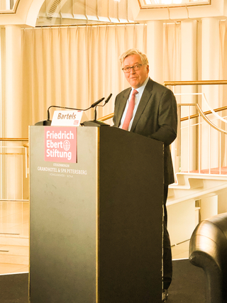 Mann im Anzug und roter Krawatte spricht am Rednerpult mit 'Friedrich-Ebert-Stiftung' und 'Bartels'-Namensschild.