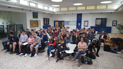 Eine große Gruppe junger Menschen sitzt in einem hellen Raum und hört einer Präsentation oder einem Vortrag zu.