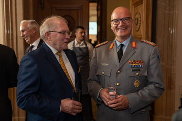 Ein Mann im Anzug spricht mit einem lachenden Militäroffizier in grauer Uniform bei einem Empfang. Beide halten Gläser.