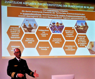 Ein deutscher Marineoffizier präsentiert eine Folie über zusätzliche Aufgaben der Bundeswehr im Inland, die diverse militärische Heimataufgaben detailliert.