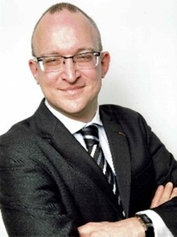 Lächelnder Mann mit Brille, glatzköpfig, im dunklen Anzug mit gestreifter Krawatte und verschränkten Armen vor weißem Hintergrund.
