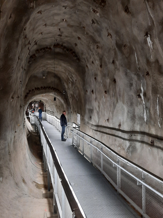 Ein Mann steht auf einem Metallsteg in einem großen, gekrümmten Betontunnel mit rauen Wänden. Weitere Personen sind in der Ferne zu sehen.