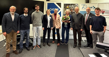 Neun Personen, darunter zwei Frauen und sieben Männer, stehen in einer Reihe vor Veranstaltungsbannern. Eine Frau hält einen Strauß.