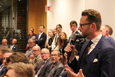 Mann im Anzug spricht ins Mikrofon auf einer Konferenz vor Publikum.
